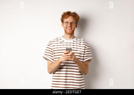 Technology and e-commerce concept. Redhead guy in glasses using mobile phone and smiling. Young man with smartphone staring happy at camera, white Stock Photo