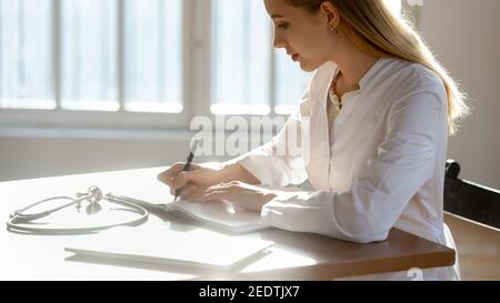 Concentrated young female doctor making records in paper copybook. Stock Photo