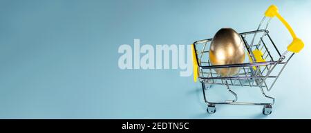 Banner with golden egg in a shopping cart. Happy Easter concept. Painted egg in yellow shopping trolley on pastel blue background. copy space Stock Photo