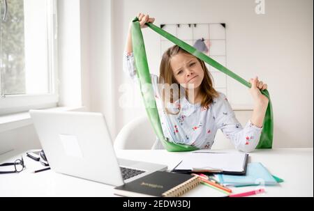 pretty blonde girl doing fitness exercises with green rubber band sitting in front of her laptop during homeschooling Stock Photo