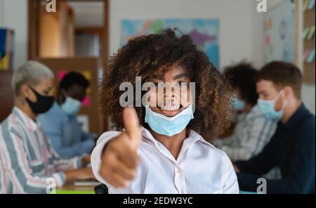 African woman thumb up wearing face mask in co-working creative space with team work - Young people working during corona virus pandemic Stock Photo