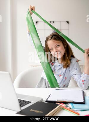 pretty blonde girl doing fitness exercises with green rubber band sitting in front of her laptop during homeschooling Stock Photo