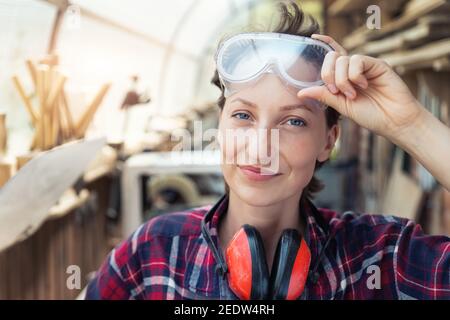 Young beautiful handy professional happy female strong carpenter portrait wearing protective goggles working in carpentry diy workshop against wood Stock Photo