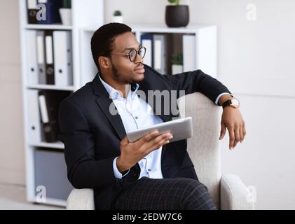 Thoughtful black businessman sitting in chair, using digital tablet Stock Photo