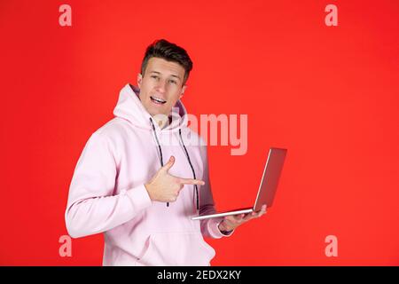 Pointing on laptop, cheerful. Caucasian man's portrait isolated on red background with copyspace. Handsome male model in street style. Concept of human emotions, facial expression, sales, ad, fashion. Stock Photo