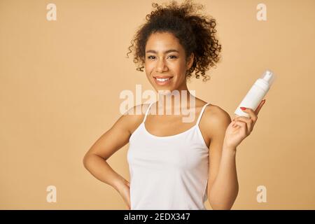 Attractive young woman smiling at camera, holding a bottle of foam facial cleanser, posing isolated over beige background. Beauty products and skin care concept Stock Photo