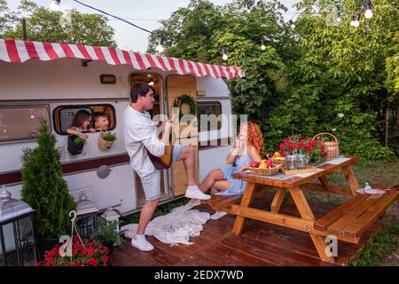Young father plays the guitar to girl. Red-haired woman mother listens to the song, laughs. Daughter and son look out the trailer window. Summer fun t Stock Photo