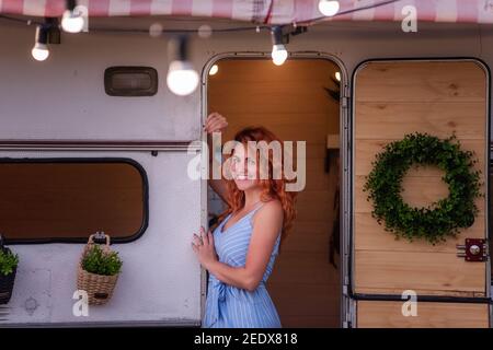A beautiful slender girl with red curly hair and a gorgeous smile in a blue sundress sits in the doorway of her trailer. A tender mother travels with Stock Photo