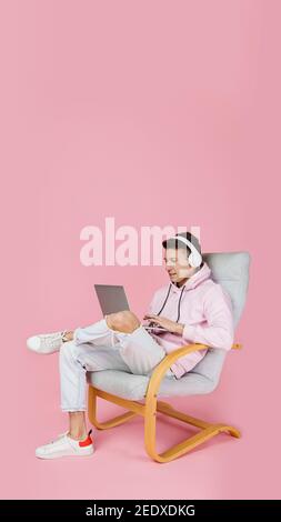 Using laptop, listening to music with headphones. Caucasian man's portrait isolated on pink background with copyspace. Handsome male model. Concept of human emotions, facial expression, sales, ad, fashion. Stock Photo