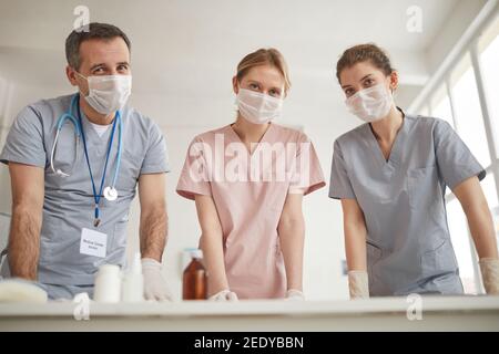 Low angle portrait of three doctors wearing masks and looking at camera while standing by desk in medical clinic Stock Photo