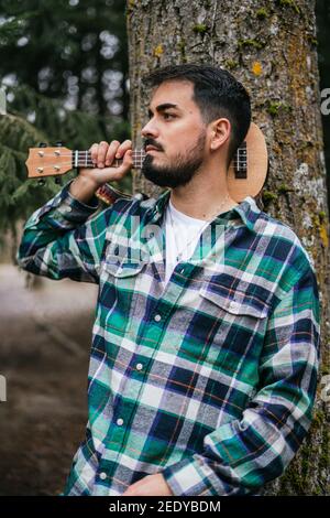 Bearded Hispanic man wearing flannel holding a ukulele while leaning against a tree Stock Photo