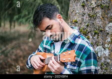 Bearded Hispanic man wearing flannel playing ukulele in the park Stock Photo