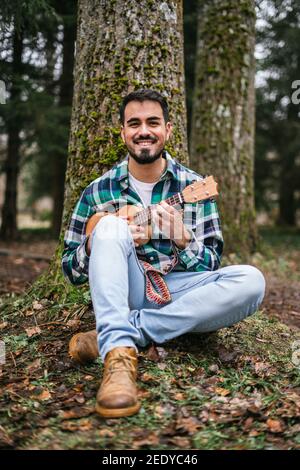 Cheerful bearded Hispanic man wearing flannel playing the ukulele while sitting under a tree Stock Photo
