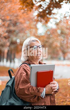 Caucasian woman holding notebook folders in her hands walking in the park. Teacher in casual clothes, glasses and with backpack. Education, studying foreign languages concept, copy space. Stock Photo