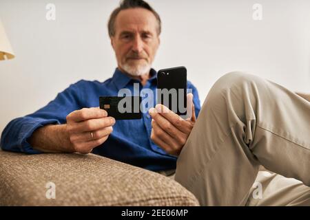 Close up of aged male hands holding a credit card and smartphone. Senior man using payment service Stock Photo