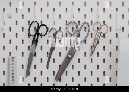 Tailor scissors in different sizes and forms hanging on hooks of perforated white wall Stock Photo