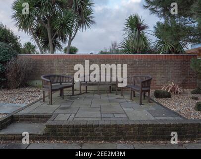 Outdoor benches seen on a raised paved area patio, with a garden wall, pebbles, bricks, plants and trees. Stock Photo