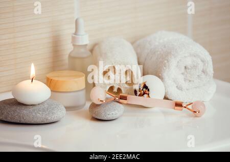 Home spa self care products in bathroom, facial massage rolling tool, moisturizing creams and oil, creamy bath bomb with dry rose petals. Stock Photo
