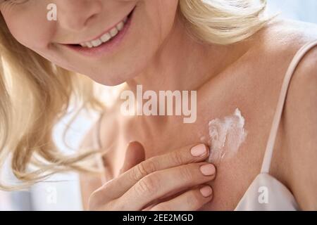 Happy middle aged woman applying cream moisturizing lotion on body skin. Stock Photo