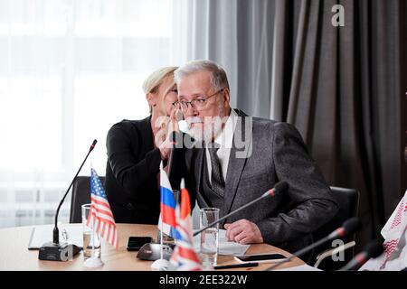 caucasian delegates sharing opinions about colleague speech, adult female talk something in the ear of senior gray bearded man in suit, discussing Stock Photo