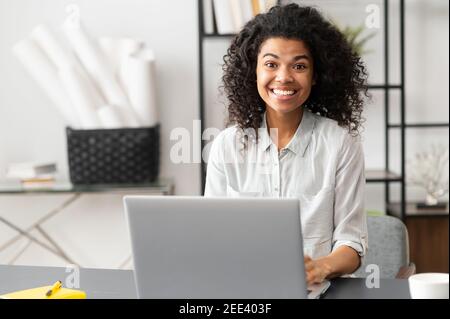 Excited young African American female office worker businesswoman with Afro hairstyle sitting at the desk with laptop, looking at the camera. Portrait Stock Photo
