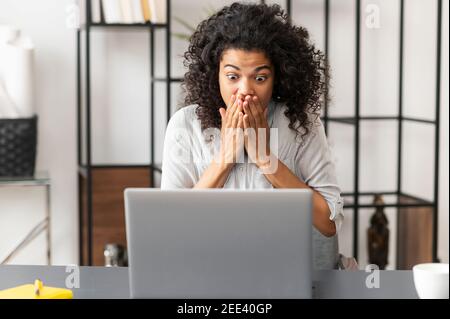 Young African American businesswoman with Afro hairstyle, eyes wide open covering mouth with hands, sitting at the desk, checking news or data online Stock Photo