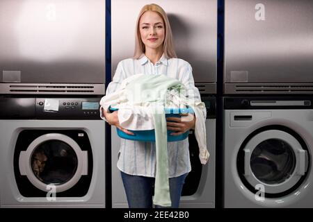 blond happy woman holding a basket of clothes to be washed in a automatic laundry, young lady stand smiling at camera. washing, cleaning, household ch Stock Photo