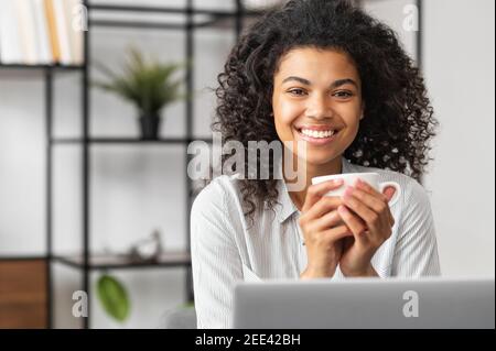 Smiling young cheerful African American woman with Afro hairstyle enjoying morning coffee while working on the laptop, sitting at the desk in the home Stock Photo