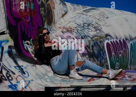 A tall blonde teenage girl wearing ripped jeans and a white tank top poses for portraits by abandoned airplanes covered in graffiti in the desert wild Stock Photo