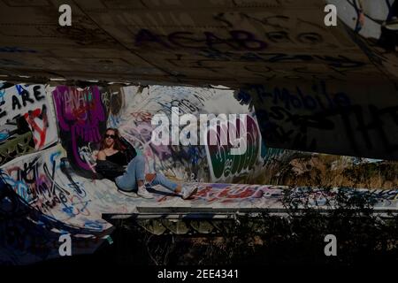 A tall blonde teenage girl wearing ripped jeans and a white tank top poses for portraits by abandoned airplanes covered in graffiti in the desert wild Stock Photo