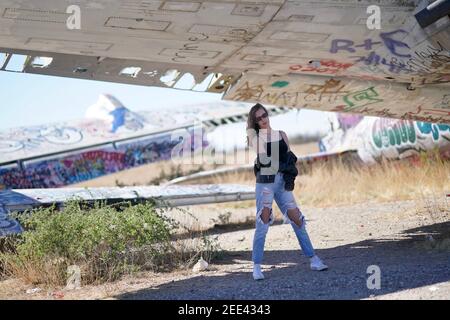 A tall blonde teenage girl wearing ripped jeans and a white tank top poses for portraits by abandoned airplanes covered in graffiti in the desert wild Stock Photo