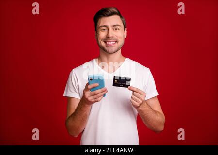 Photo portrait of young man keeping mobile phone demonstrating plastic bank card isolated vivid red color background Stock Photo