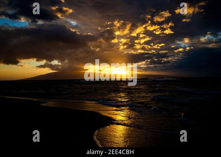 Golden sunset seascape with light rays beaming from beneath clouds as sun sets behind tropical island. Stock Photo
