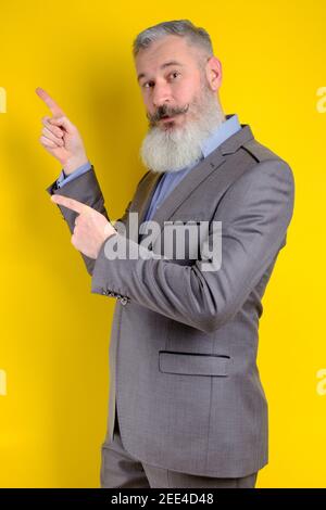 Studio portrait mature businessman dressed in gray suit points aside, I choose you this, yellow background Stock Photo