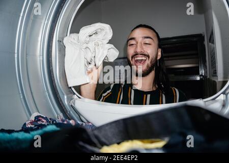 stylish young man with long hair is washing clothes in a laundromat, throwing clothes into the washing machine. dryer, living alone, washing clothes a Stock Photo