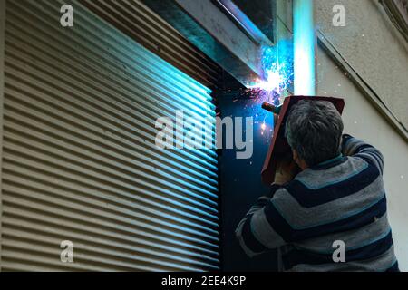 A middle aged man welds in the street. Welder worker. Man holding welding helmet during the weld. Man repairing the shutter of a shop. Stock Photo