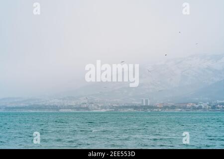 Gelendzhik city in winter in snowy weather, mountains covered with snow, waves and wind on sea bay water surface, no people walking in city. Stock Photo