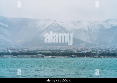 Gelendzhik city in winter in snowy weather, mountains covered with snow, waves and wind on sea bay water surface, no people walking in city. Stock Photo