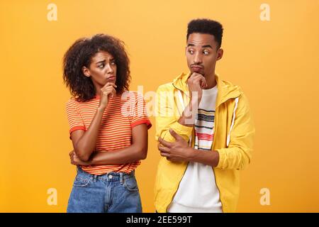 Silly confused girl standing gloomy and worried pursing lips and frowning holding hand on chin looking at boyfriend confused while guy glancing at Stock Photo