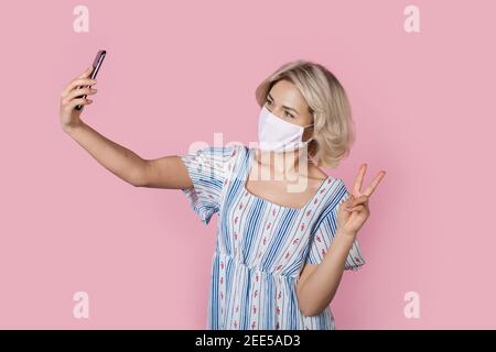 Adorable blonde lady in medical mask in gesturing the peace sign while taking a selfie on a pink wall Stock Photo