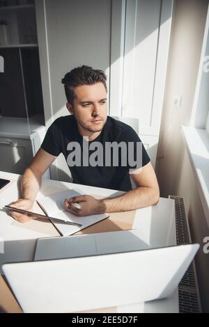 Caucasian man is working remotely from home using a laptop and thinking about something near the window Stock Photo