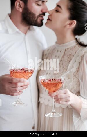 Happy romantic young caucasian couple celebrating, hugging, holding glasses of sparkling champagne cocktail with rose petals and glass drinking straws. Wedding anniversary, Valentines Day concept Stock Photo