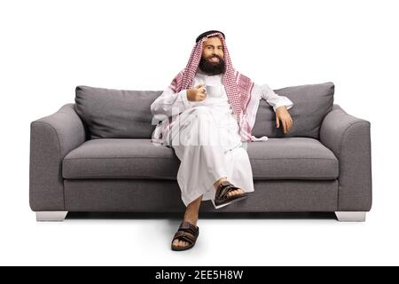 Saudi arab man sitting on a gray sofa and drinking a cup of tea isolated on white background Stock Photo