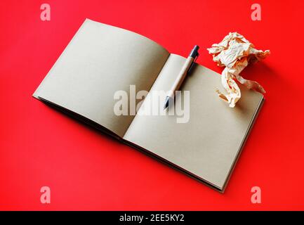 Opened book with blank craft paper pages, pen and crumpled paper on red background. Stock Photo