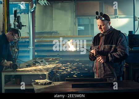 Handyman preparing his autogenous welding tool for work while his colleague can be noticed in the back working on his grinding task. Stock Photo