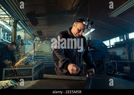 Welder working with autogenous welding tool in workshop while his colleague can be noticed in the back grinding. Stock Photo