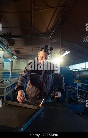 Welder working with autogenous welding tool in workshop while his colleague can be noticed in the back grinding. Stock Photo