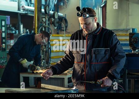 Welder working with autogenous welding tool in workshop while his colleague can be noticed in the back grinding. Stock Photo