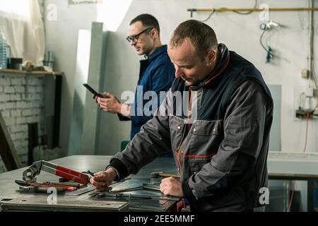 A tinsmith is measuring on a piece of the tin in his workshop and he's taking instruction from a colleague engineer who is holding tablet. Stock Photo