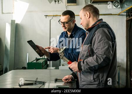A tinsmith and his fellow colleague engineer are working together on a work task in their workshop, while the engineer is using tablet. Stock Photo